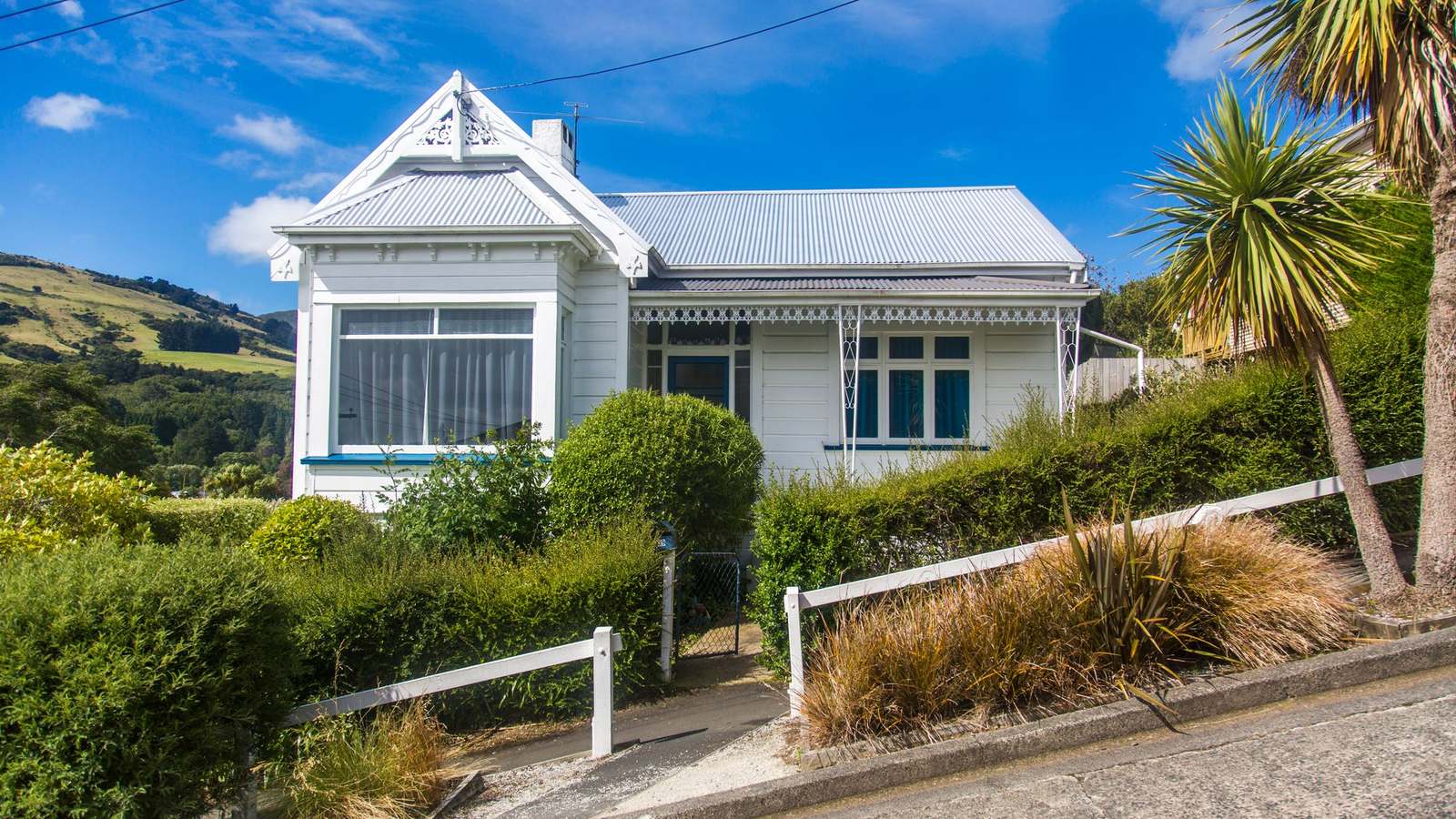 A white weatherboard villa on a steep Dunedin street, with cabbage trees and flax