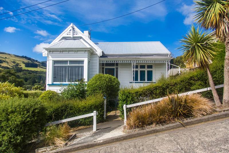 A white weatherboard villa on a steep Dunedin street, with cabbage trees and flax