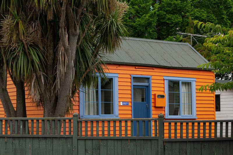 A tiny orange weatherboard cottage with a picket fence and a cabbage tree in Dunedin