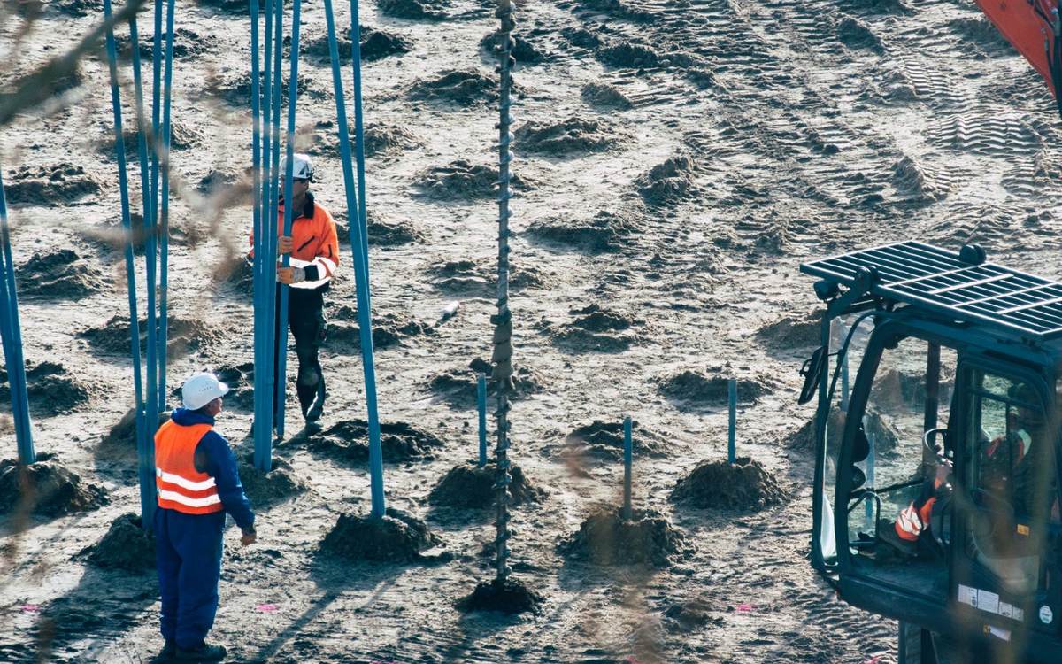 An excavator with a drive head beside rows of steel piles, with two workers