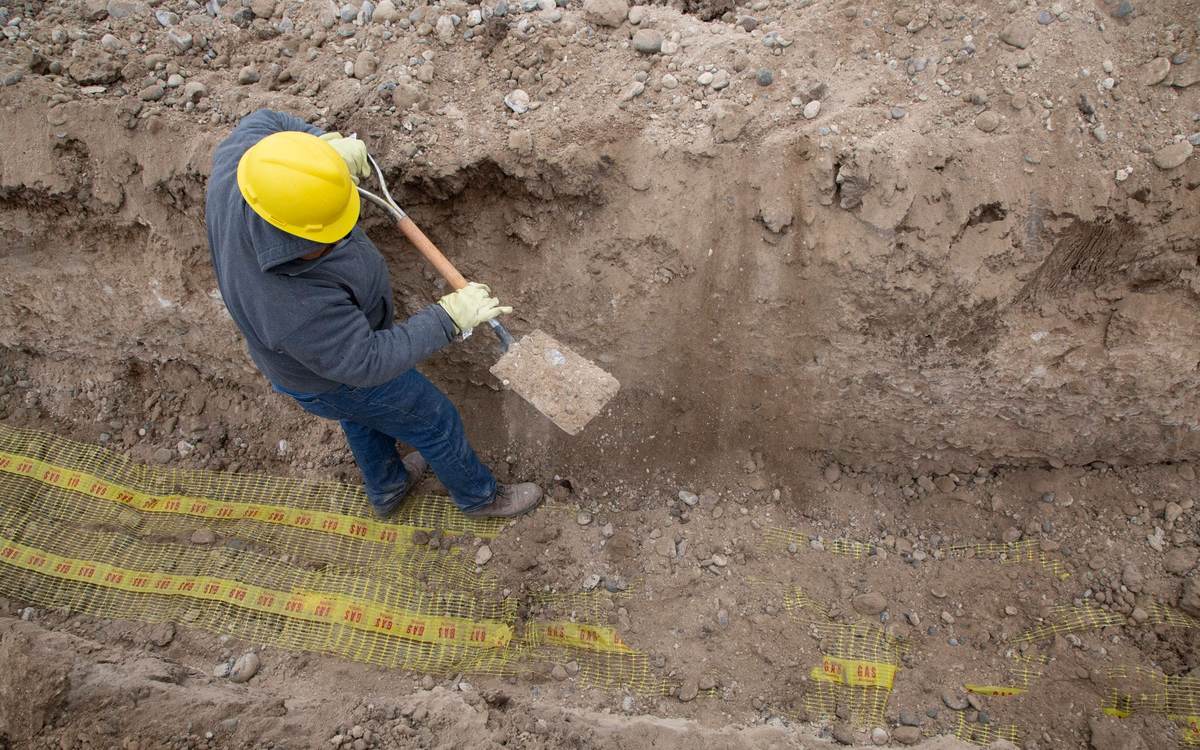 A worker digging a trench that shows layers of soil and gravel